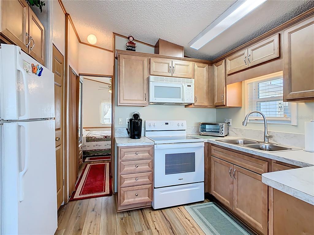 A kitchen with light wood cabinets, white appliances, and a view into an adjacent bedroom — 9000 US HWY 192 UNIT 311, CLERMONT, FL
