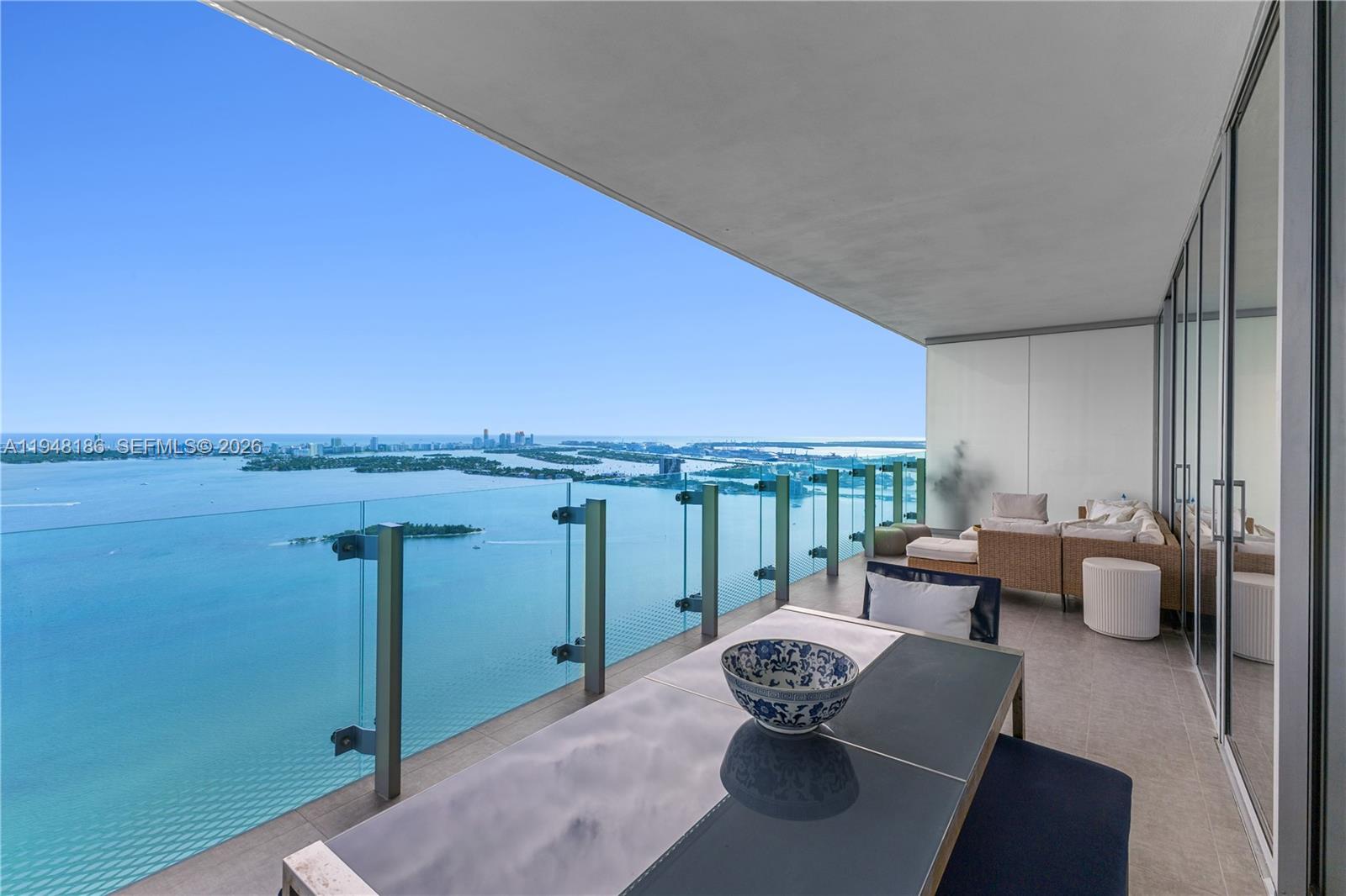 A high-rise balcony features a glass railing, outdoor dining set, and lounge furniture overlooking water — 700 NE 26th Ter # 4701, Miami FL 33137, MIAMI, FL