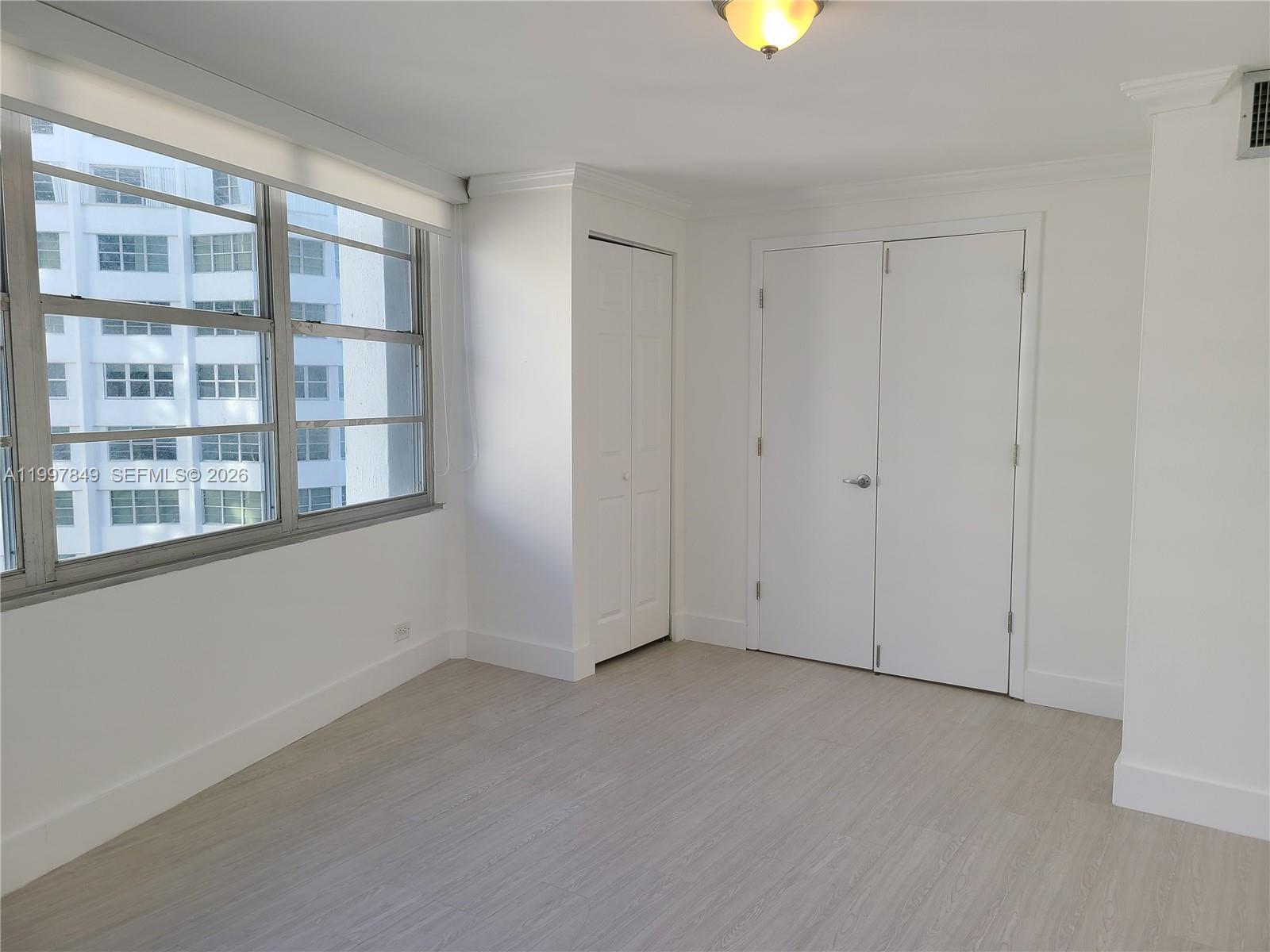 An empty room features white walls, light wood-look flooring, two sets of closets, and windows — 999 Brickell Bay Dr # 1207, Miami FL 33131, MIAMI, FL