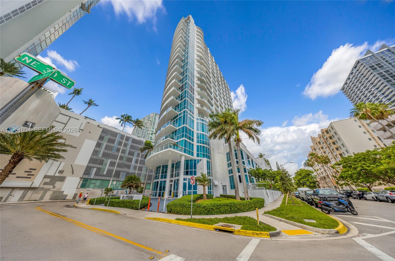 A modern glass-facade high-rise condominium building stands on a street corner with palm trees — 480 NE 30th St # 1402, Miami FL 33137, MIAMI, FL