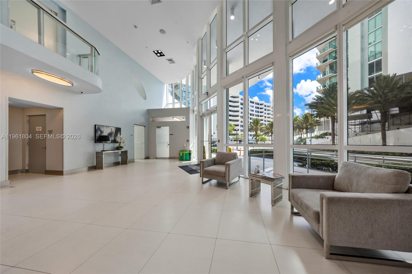 A bright, high-ceilinged lobby features white tile floors, floor-to-ceiling windows, and two gray armchairs — 480 NE 30th St # 1402, Miami FL 33137, MIAMI, FL