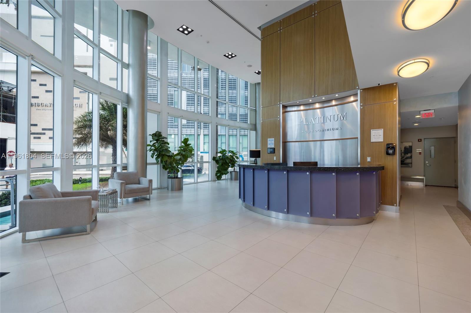 A modern condominium lobby features a curved reception desk, floor-to-ceiling windows, and tile flooring — 480 NE 30th St # 1402, Miami FL 33137, MIAMI, FL