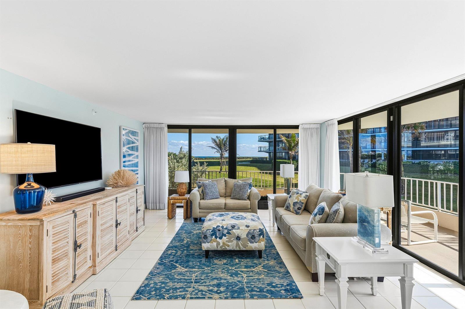A living room features a blue patterned rug, beige sofas, and large windows overlooking greenery — 3400 S Ocean Boulevard, Palm Beach FL 33480, PALM BEACH, FL