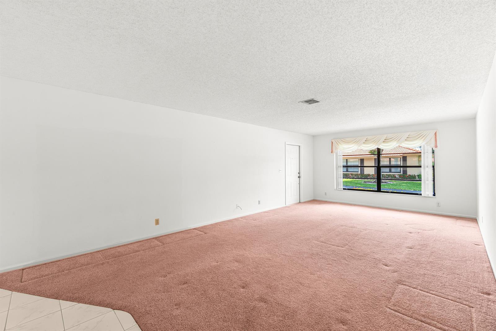 An empty living room features pink wall-to-wall carpeting, white walls, and a large window — 4655 Mahoe Tree Place # A, Boynton Beach FL 33436, BOYNTON BEACH, FL