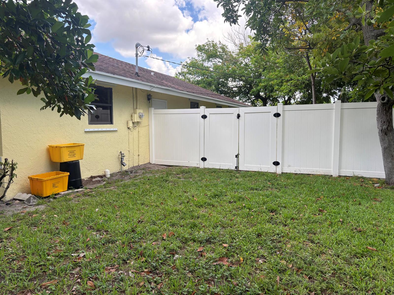 A yellow house exterior features a white vinyl privacy fence and a grassy backyard — 1563 40th Street, West Palm Beach FL 33407, WEST PALM BEACH, FL