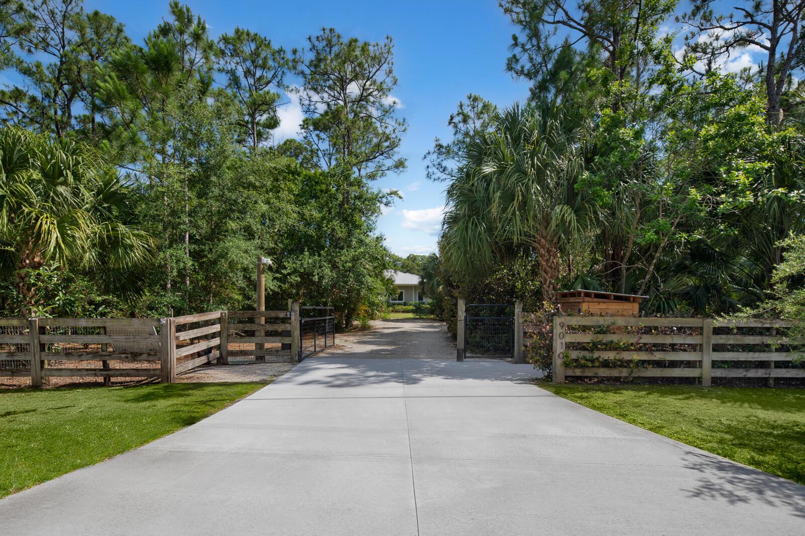 A concrete driveway leads through a wooden gate toward a house surrounded by lush trees — 9010 Valley Oak Place, Jupiter FL 33478, JUPITER, FL