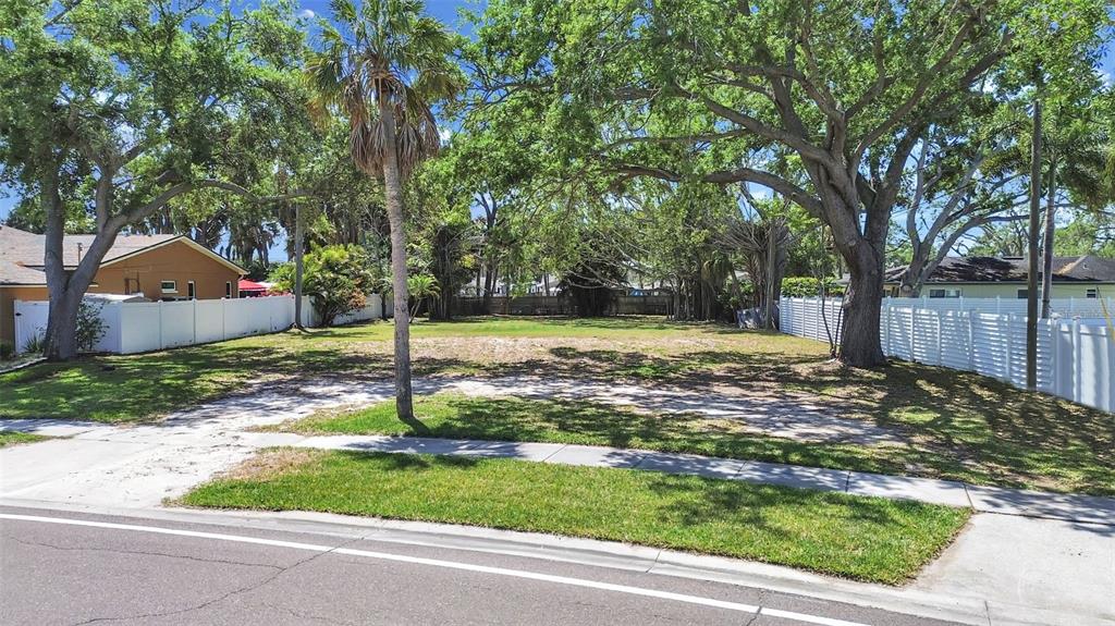 A vacant, grassy residential lot sits between two white fenced properties under large shade trees — 5438 BAYOU GRANDE BOULEVARD NE, ST PETERSBURG, FL