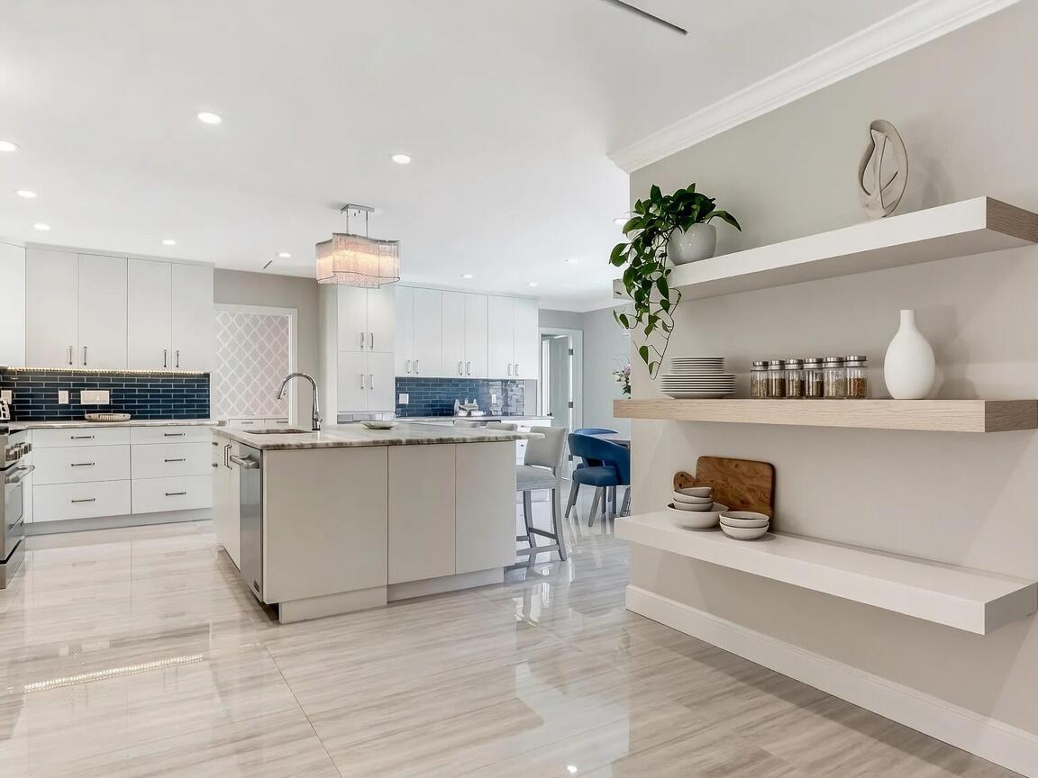 A modern kitchen features white cabinetry, a central island, blue tile backsplash, and floating shelves — 831 Dover Street, Boca Raton FL 33487, BOCA RATON, FL