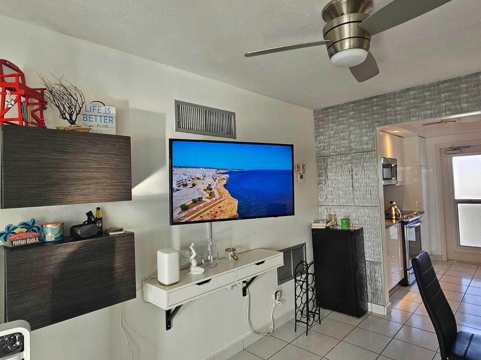 A living room with a wall-mounted television, floating white shelf, and an adjacent kitchen area — 505 N Riverside Drive # 12a, Pompano Beach FL 33062, POMPANO BEACH, FL