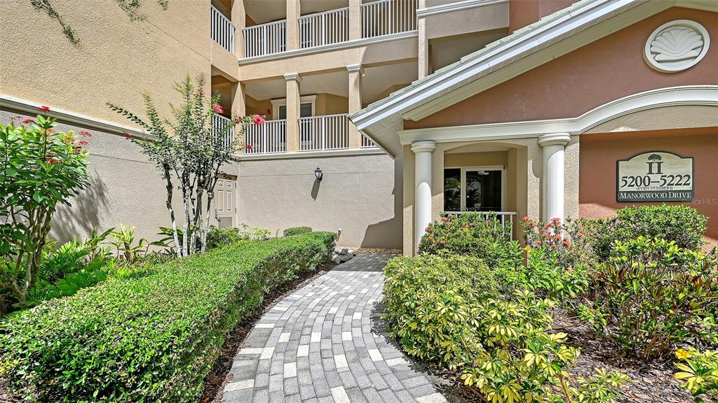 A paved walkway leads to the entrance of a multi-story building with landscaped bushes — 5222 MANORWOOD DRIVE UNIT 4A, SARASOTA, FL