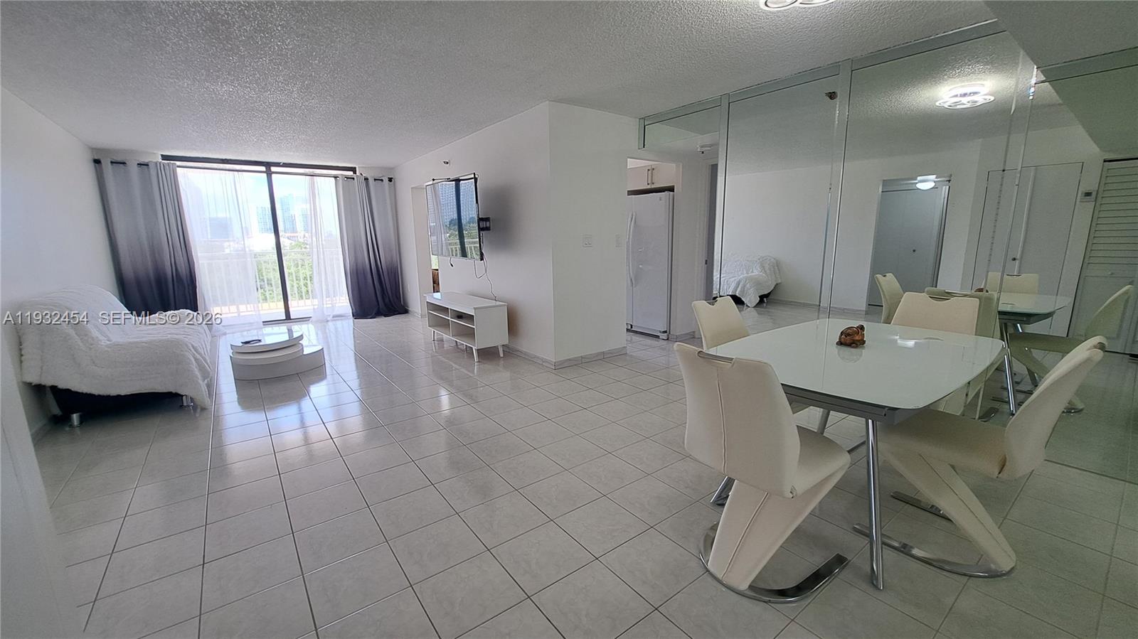 A bright living and dining area features white tile floors, mirrored walls, and balcony access — SUNNY ISLES BEACH, FL