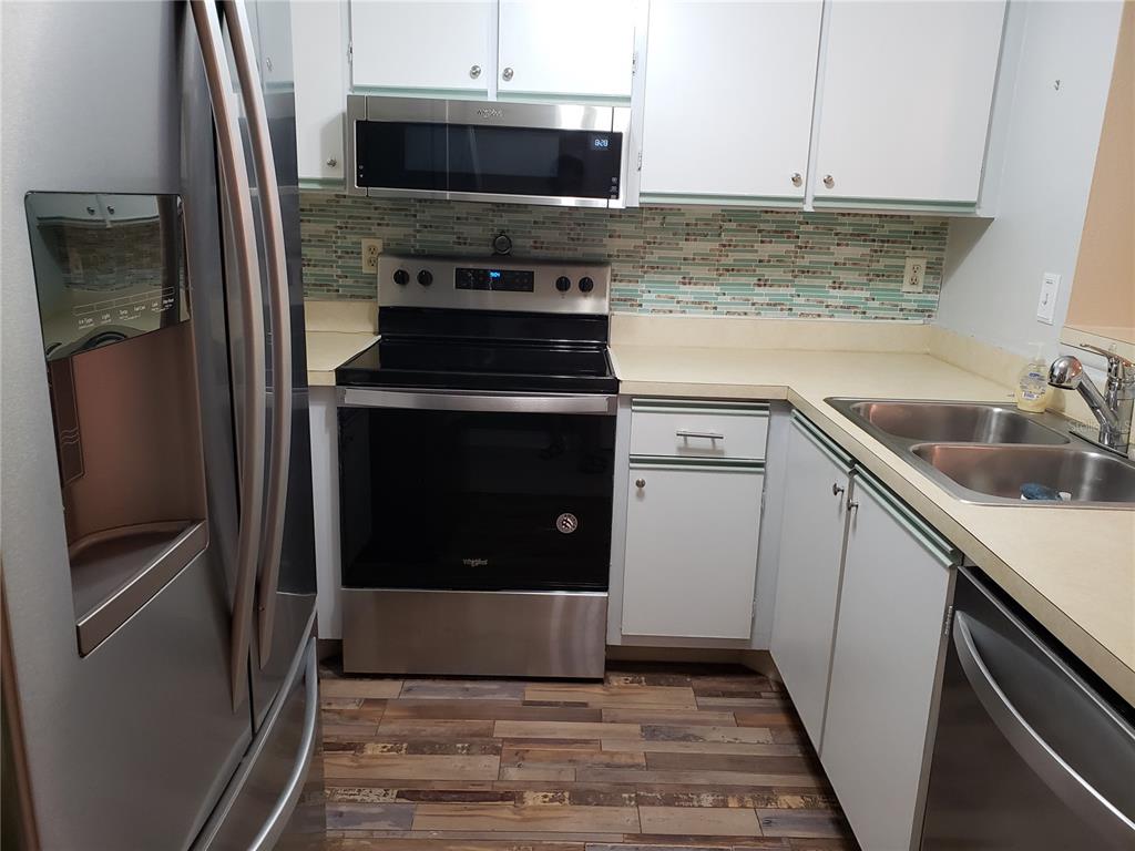 A kitchen features stainless steel appliances, white cabinets, a tiled backsplash, and wood-look flooring — 19505 QUESADA UNIT OO103, PORT CHARLOTTE, FL