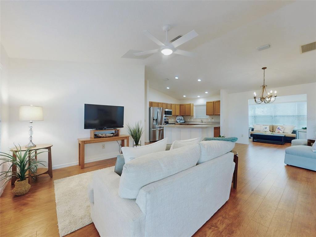An open-concept living room features a white sofa, wood flooring, and a kitchen beyond — 1521 HOLLYBERRY PLACE, THE VILLAGES, FL