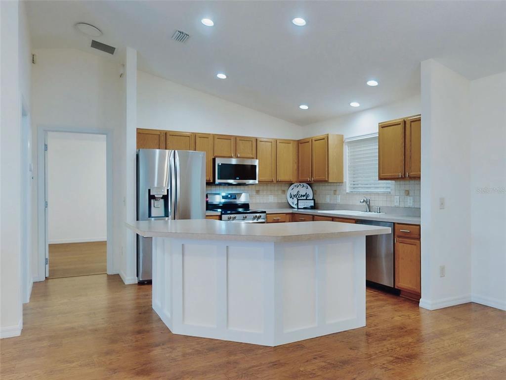 A kitchen features wood cabinets, stainless steel appliances, a white island, and wood-look flooring — 1521 HOLLYBERRY PLACE, THE VILLAGES, FL