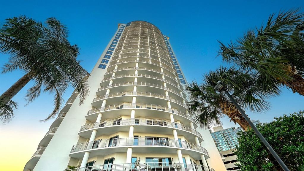 A low-angle exterior view of a tall, curved residential high-rise building flanked by palm trees — 449 S 12TH STREET UNIT 1406, TAMPA, FL