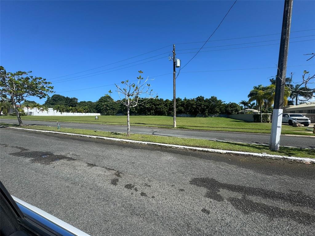 A vacant, grassy residential lot sits across a paved road under a clear blue sky — Hacienda El Molino A-10 CALLE PASEO MADRID, VEGA ALTA, PR