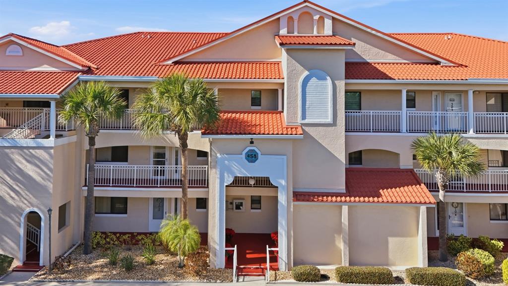 A multi-story residential building features a red tile roof, beige stucco walls, and palm trees — 458 BOUCHELLE DRIVE UNIT 203, NEW SMYRNA BEACH, FL