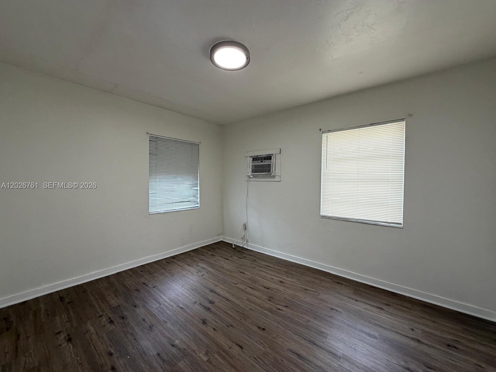 An empty room features white walls, dark wood-look flooring, two windows, and a wall-mounted air conditioner — 541 SW 7th St # 3, Miami FL 33130, MIAMI, FL
