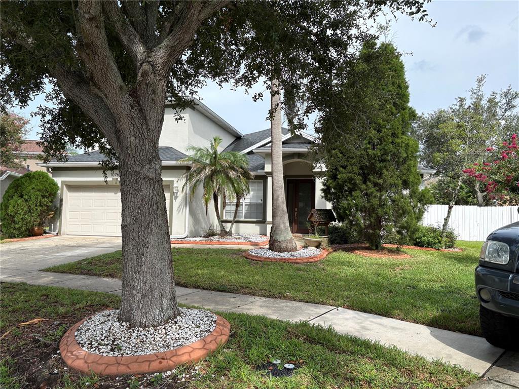 A single-story house with a garage, a large front tree, and landscaped garden beds — 10761 BANFIELD DRIVE, RIVERVIEW, FL