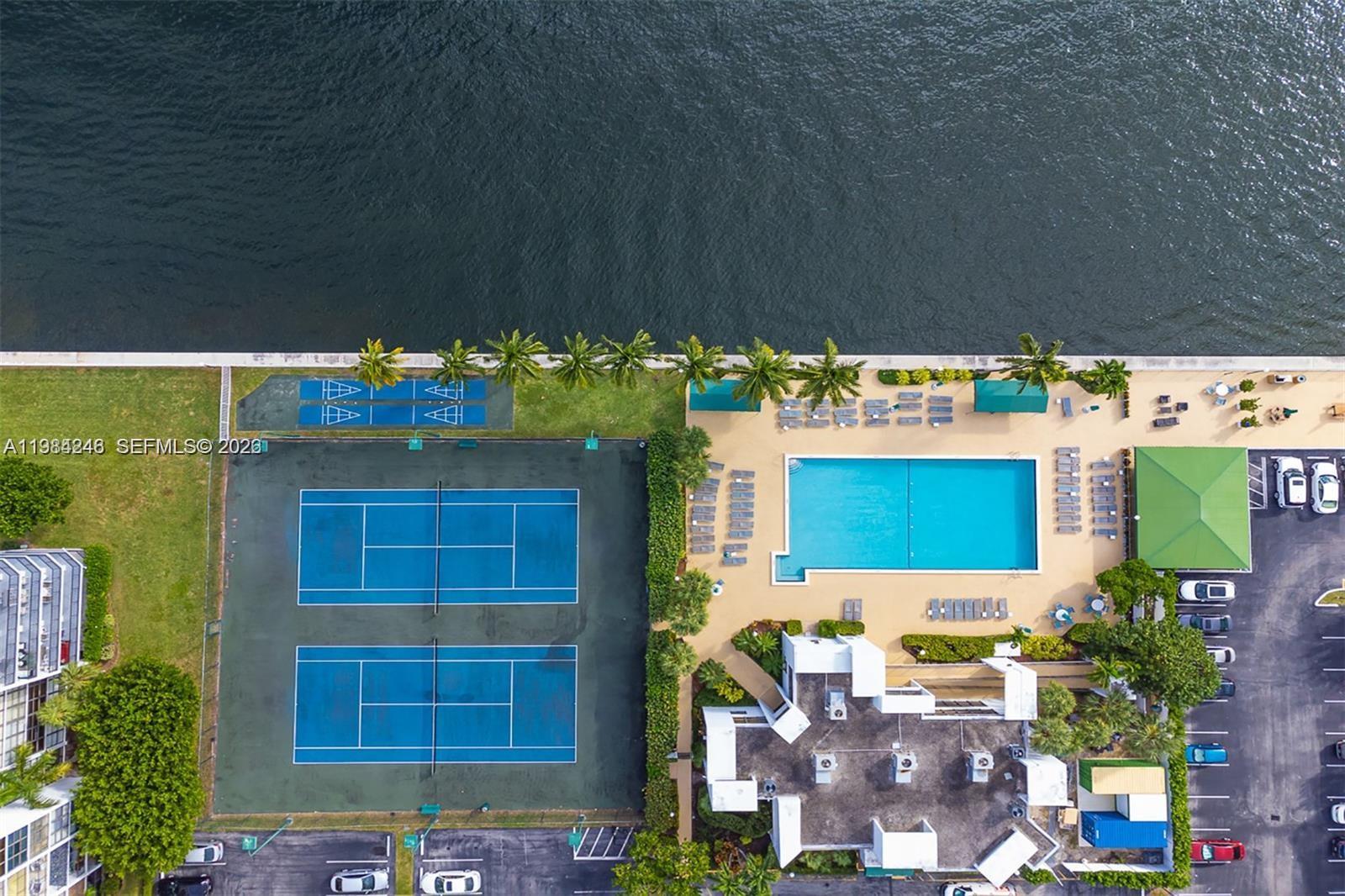 An aerial view shows tennis courts, a swimming pool, and a parking lot by water — 800 Parkview Dr # 527, Hallandale Beach FL 33009, HALLANDALE BEACH, FL