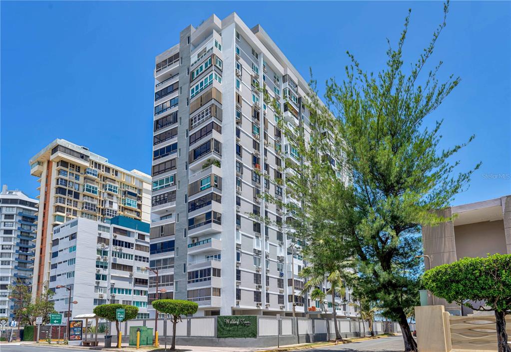 A tall residential apartment building stands under a clear blue sky next to a tree — 4123 ISLA VERDE AVENUE UNIT 1106, CAROLINA, PR