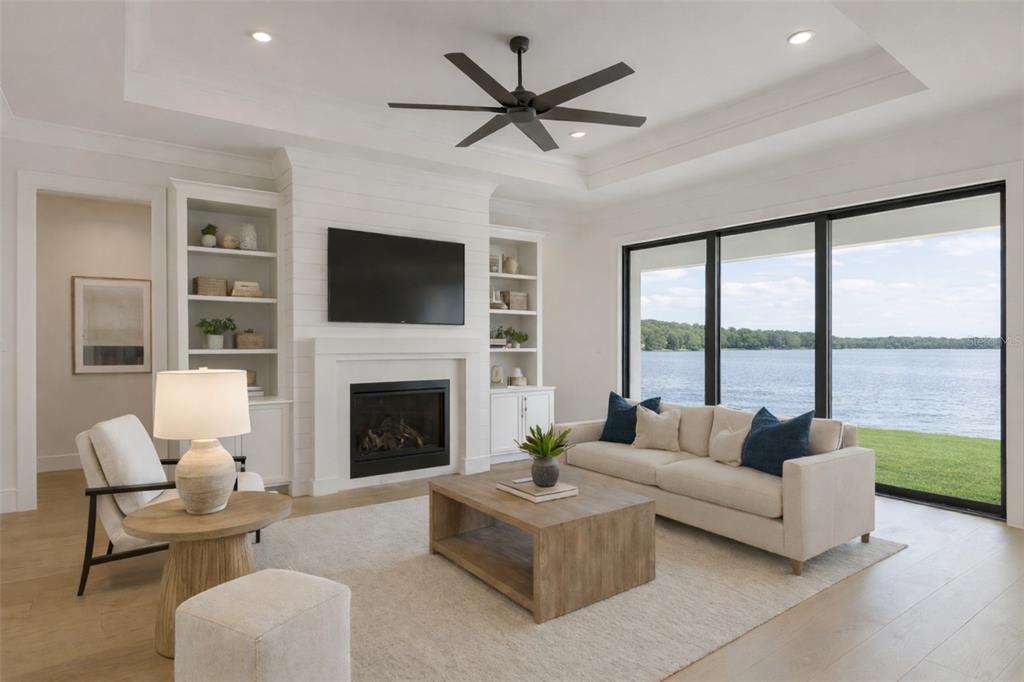 A living room features a fireplace, built-in shelving, a ceiling fan, and large lake-view windows — 6043 BAYOU GRANDE BOULEVARD NE, ST PETERSBURG, FL