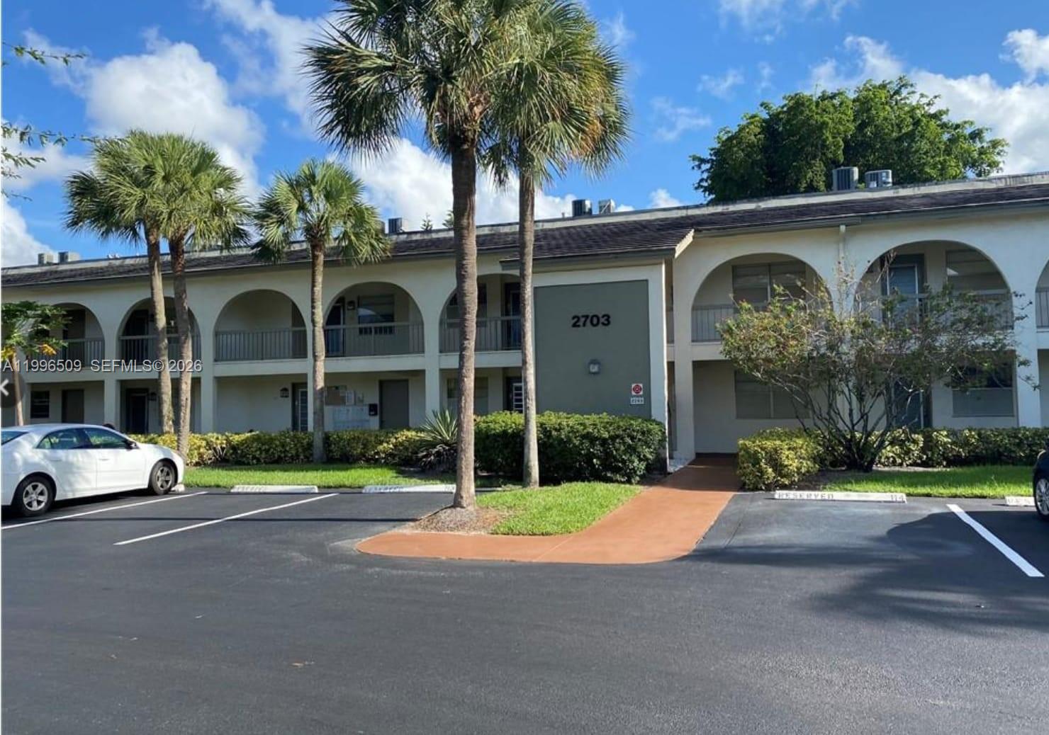 A two-story apartment building with arched balconies and a parking lot in the foreground — 2703 Nassau Bnd # E2, Coconut Creek FL 33066, COCONUT CREEK, FL