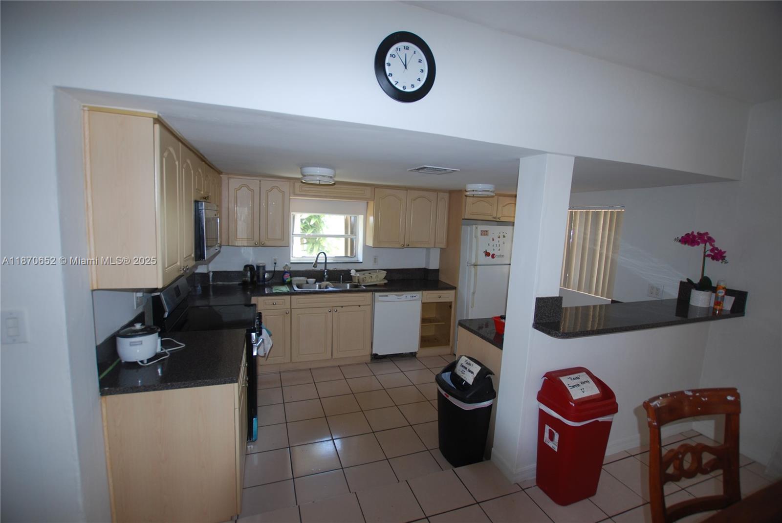 A kitchen features light wood cabinets, dark countertops, tile flooring, and a wall-mounted clock — 1801 NE 176th St, North Miami Beach FL 33162, NORTH MIAMI BEACH, FL