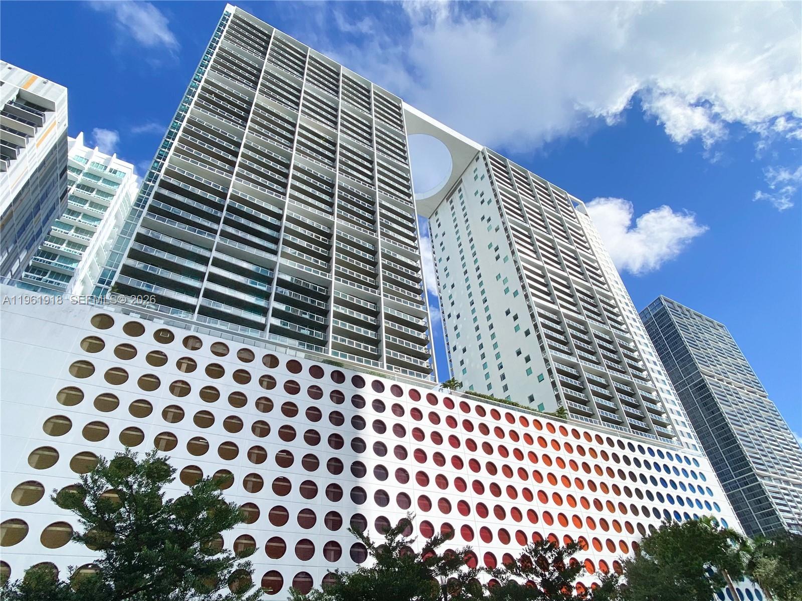 A low-angle view shows a modern high-rise building with a distinctive circular cutout and patterned facade — 55 SE 6 ST # 1207, Miami FL 33131, MIAMI, FL