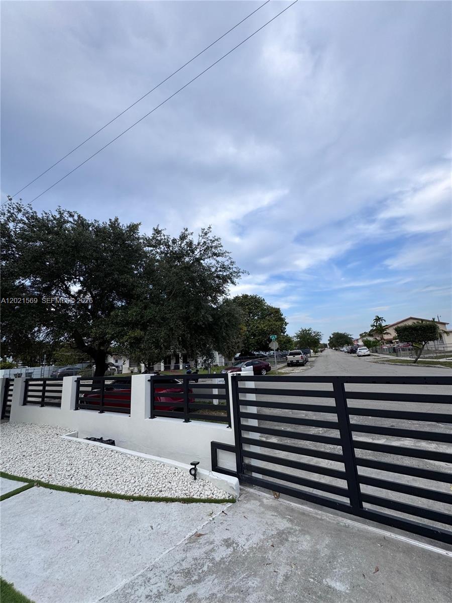 A black metal gate and white wall enclose a property with a large tree — 1120 E 6th Ct, Hialeah FL 33010, HIALEAH, FL