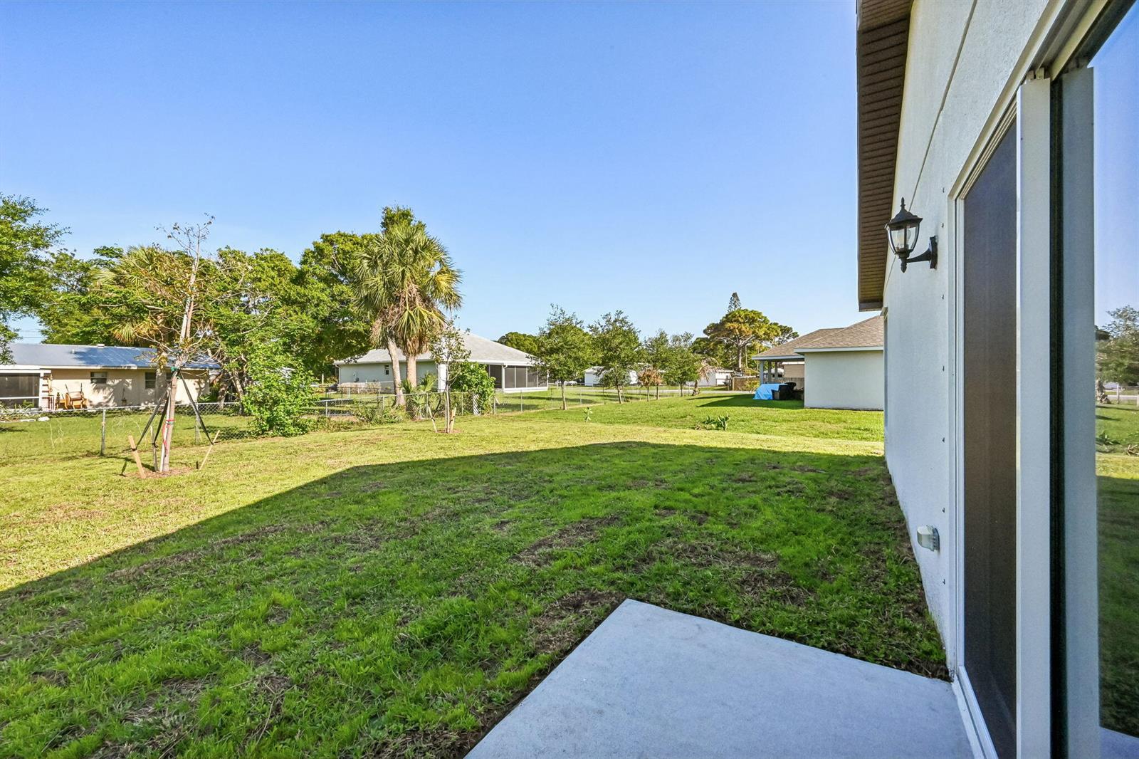 A concrete patio overlooks a grassy backyard with trees and neighboring houses under a clear sky — 5606 Seagrape Drive, Fort Pierce FL 34982, FORT PIERCE, FL