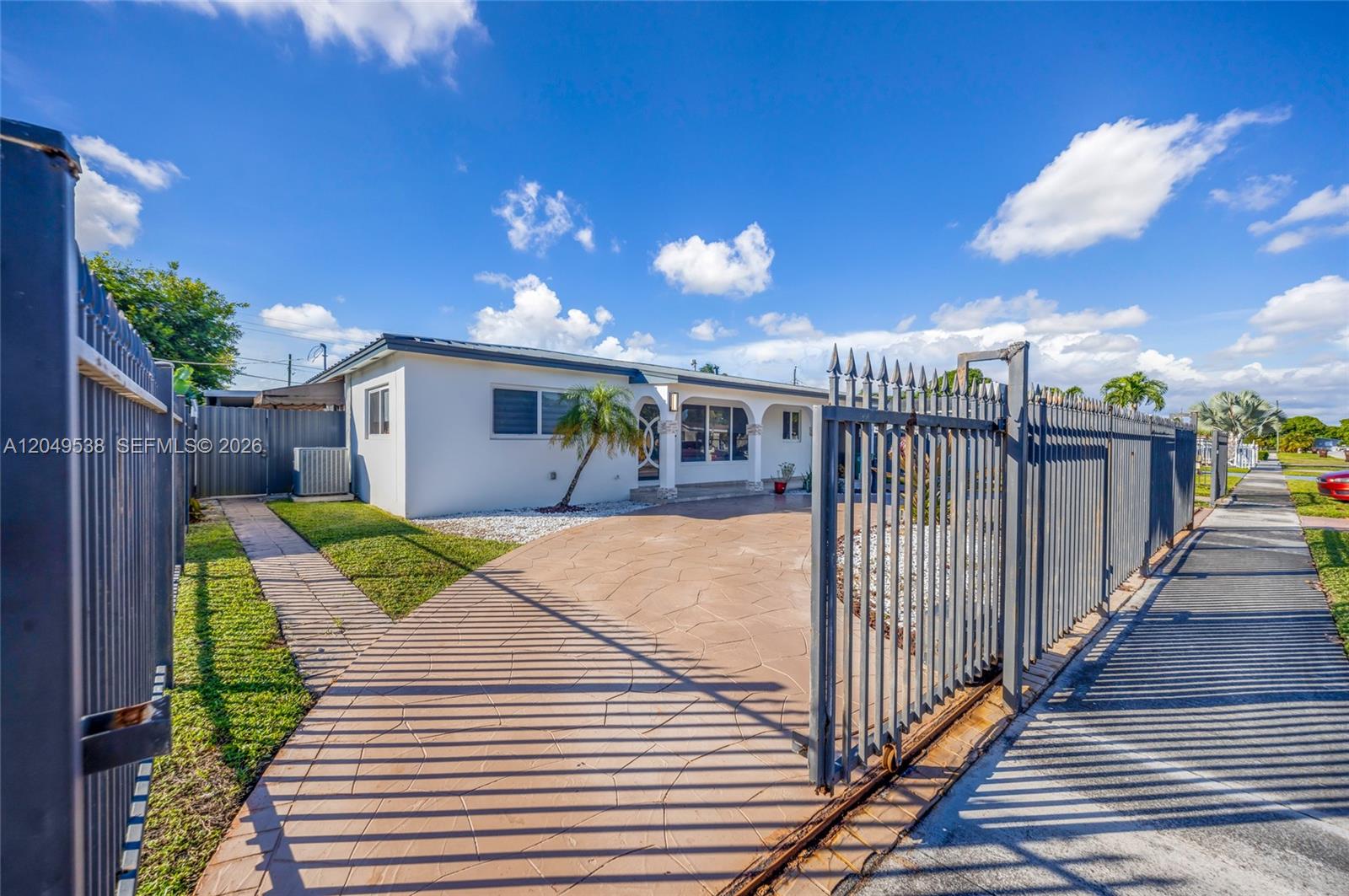 A single-story house with a paved driveway and metal gate under a blue sky — 490 W 33rd Pl, Hialeah FL 33012, HIALEAH, FL