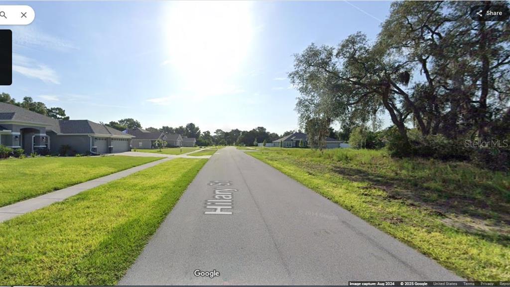 A paved street runs between a residential house on the left and a wooded lot — HILARY STREET, SPRING HILL, FL