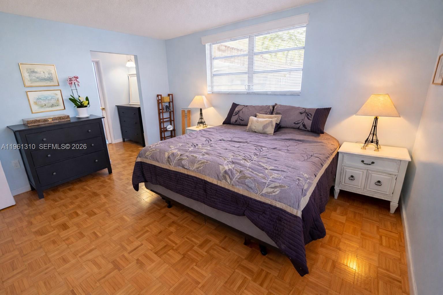 A bedroom features a bed with a purple floral quilt, parquet flooring, and light blue walls — 16750 NE 14th Ave # 202, Miami FL 33162, MIAMI, FL