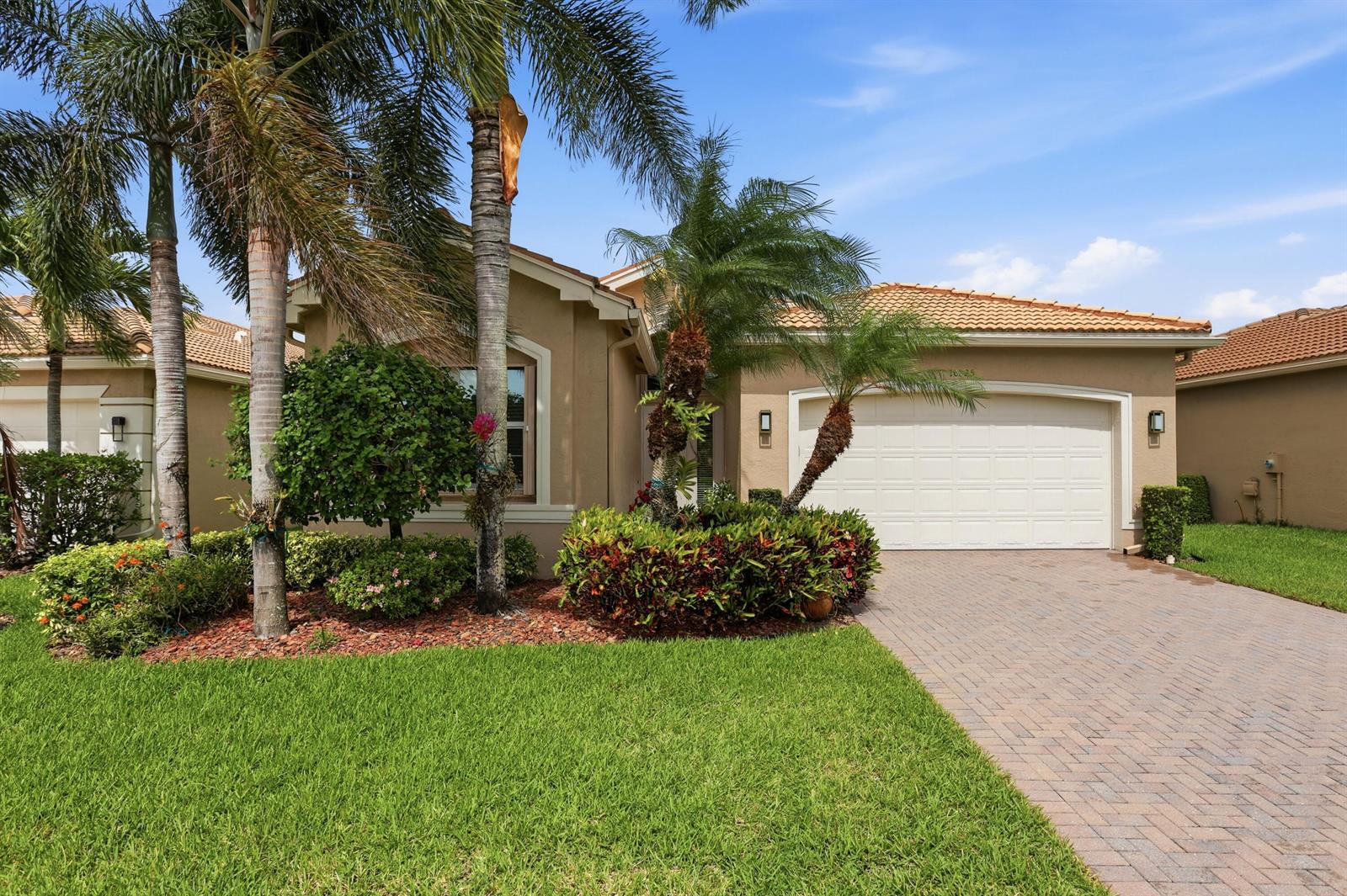 The exterior of a single-story tan house features a two-car garage and palm trees — 10825 Leaf Bridge Way, Boynton Beach FL 33473, BOYNTON BEACH, FL