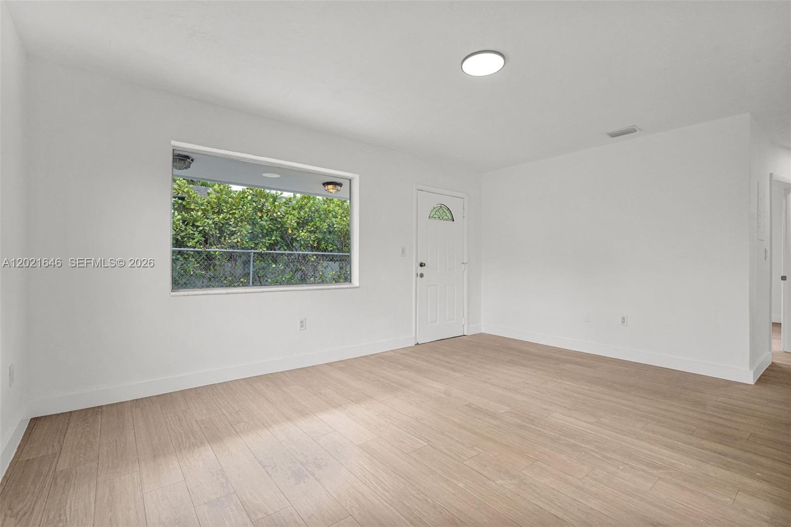A bright, empty room features white walls, light wood-look flooring, and a large window — 151 NW 42nd St # 1, Miami FL 33127, MIAMI, FL