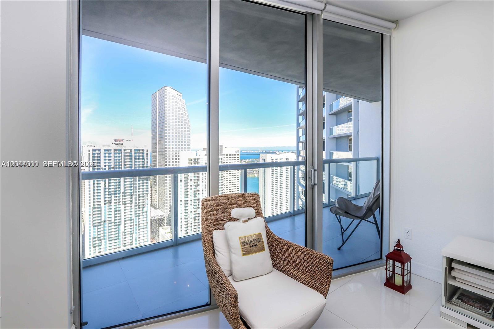 A wicker chair with a Chanel pillow sits before floor-to-ceiling windows overlooking a city skyline — 475 Brickell Ave # 4612, Miami FL 33131, MIAMI, FL