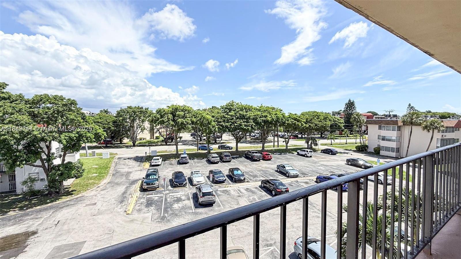 A balcony overlooks a parking lot with several cars and surrounding trees under a blue sky — 1450 NE 191st St # 404, Miami FL 33179, MIAMI, FL