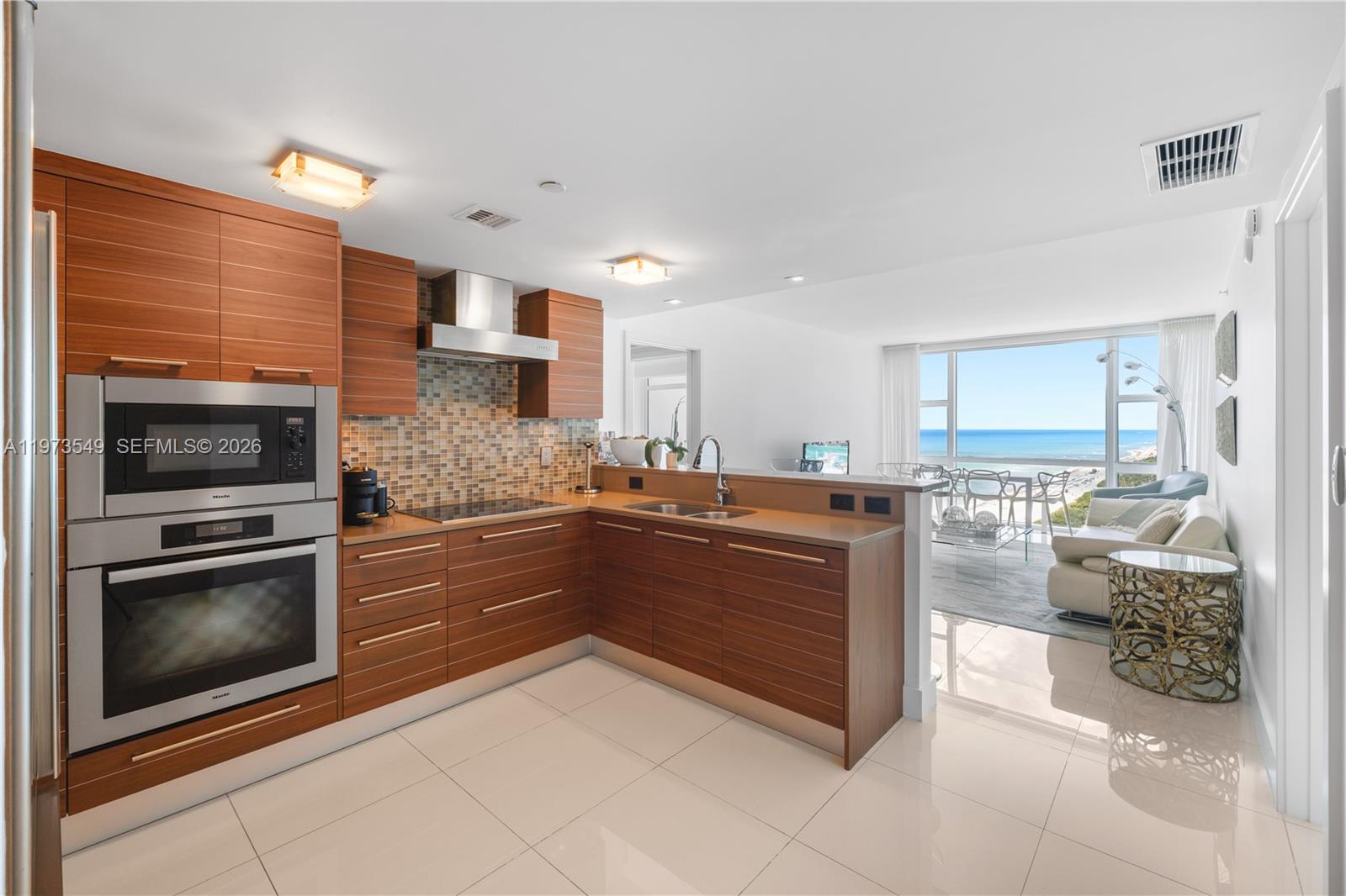 A modern kitchen with wood cabinets and stainless steel appliances overlooks a bright living area — 6801 Collins Ave, Miami Beach FL 33141, MIAMI BEACH, FL
