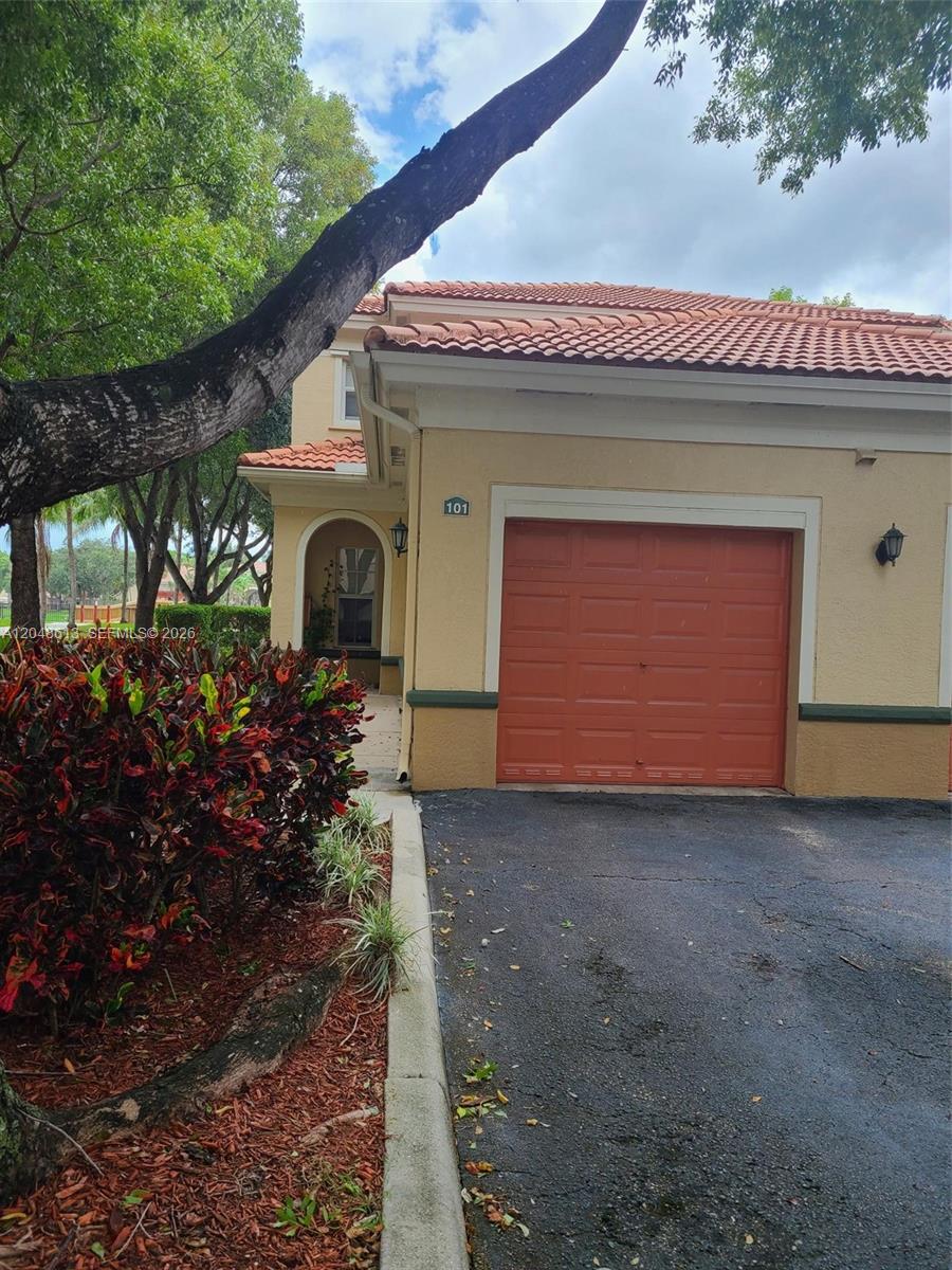 A tan house exterior features a red garage door, a tiled roof, and landscaping — 2541 Centergate Dr # 101, Miramar FL 33025, MIRAMAR, FL
