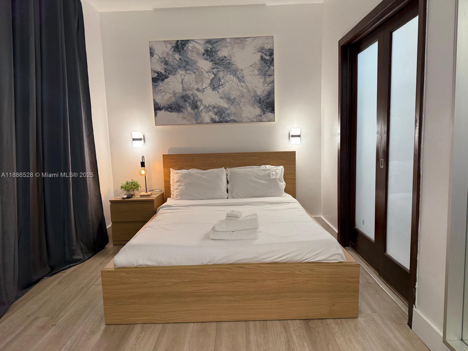 A bedroom features a wooden platform bed, a nightstand, wall sconces, and abstract wall art — 1036 Ocean Dr # B303, Miami Beach FL 33139, MIAMI BEACH, FL
