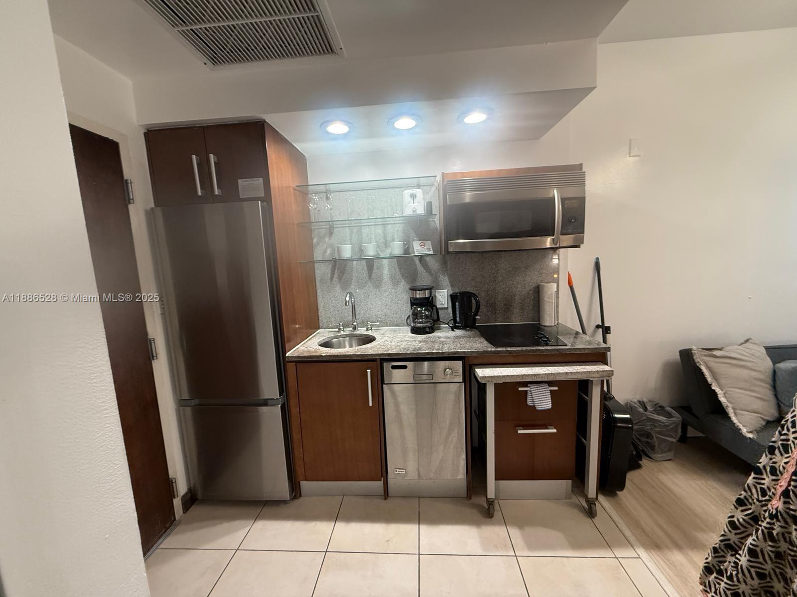 A compact kitchen features stainless steel appliances, wood cabinetry, granite countertops, and glass shelving — 1036 Ocean Dr # B303, Miami Beach FL 33139, MIAMI BEACH, FL