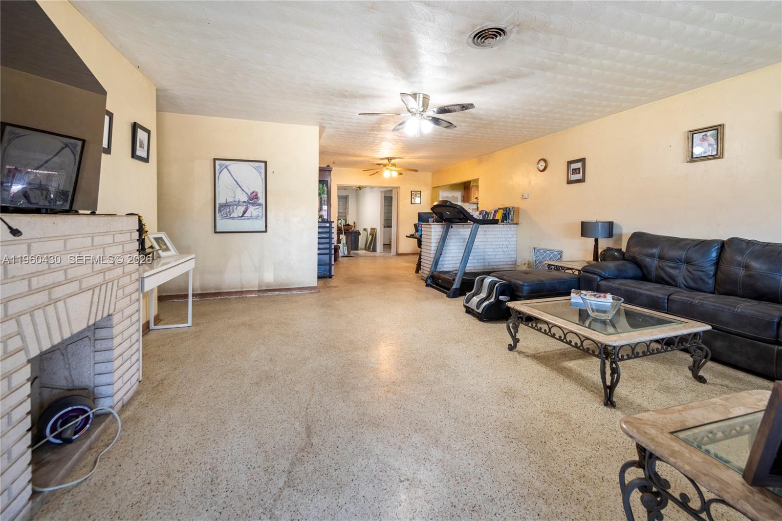 A living room features a brick fireplace, terrazzo flooring, and a treadmill near the kitchen — 600 SW 11th Ave, Hallandale Beach FL 33009, HALLANDALE BEACH, FL