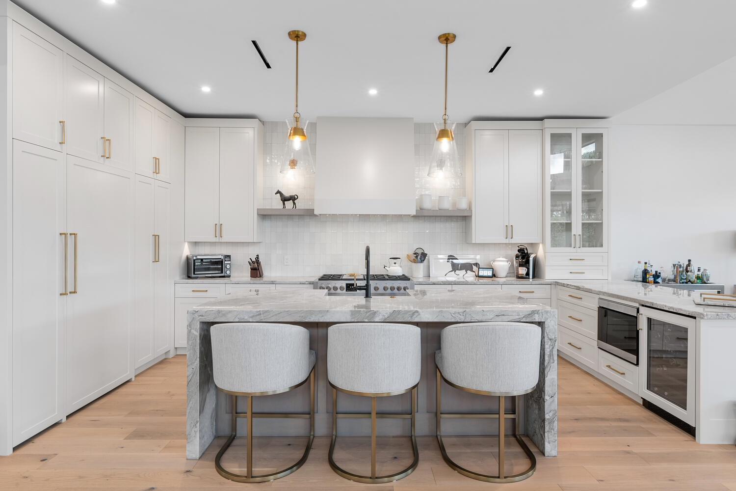 A modern kitchen features white cabinetry, a marble-topped island with three stools, and gold lighting — 12618 Mallet Circle, Wellington FL 33414, WELLINGTON, FL