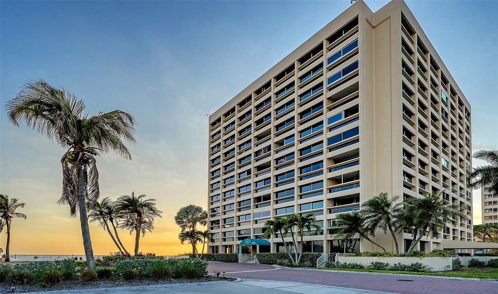 A tall, beige multi-story apartment building stands near a beach with palm trees at sunset — 5966 MIDNIGHT PASS ROAD UNIT G-67, SARASOTA, FL