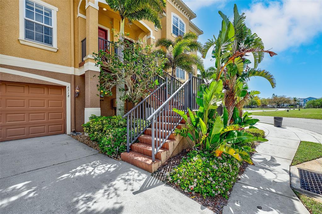A tan multi-story building features a garage, an outdoor staircase, and lush tropical landscaping — 6518 SAND SHORE LANE, NEW PORT RICHEY, FL