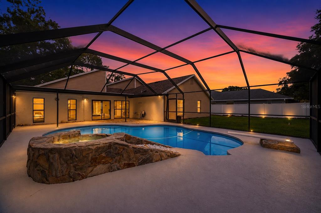 A screened-in pool and hot tub with stone accents sit behind a house at sunset — 14207 ASHBURN PLACE, TAMPA, FL