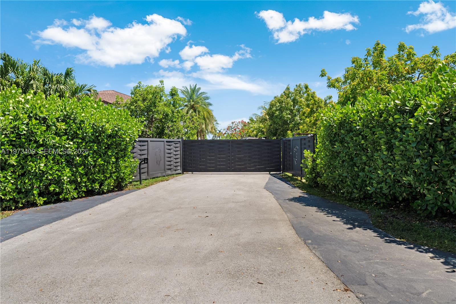 A paved driveway leads to a closed black metal gate flanked by tall green hedges — 13050 SW 26th St, Miami FL 33175, MIAMI, FL
