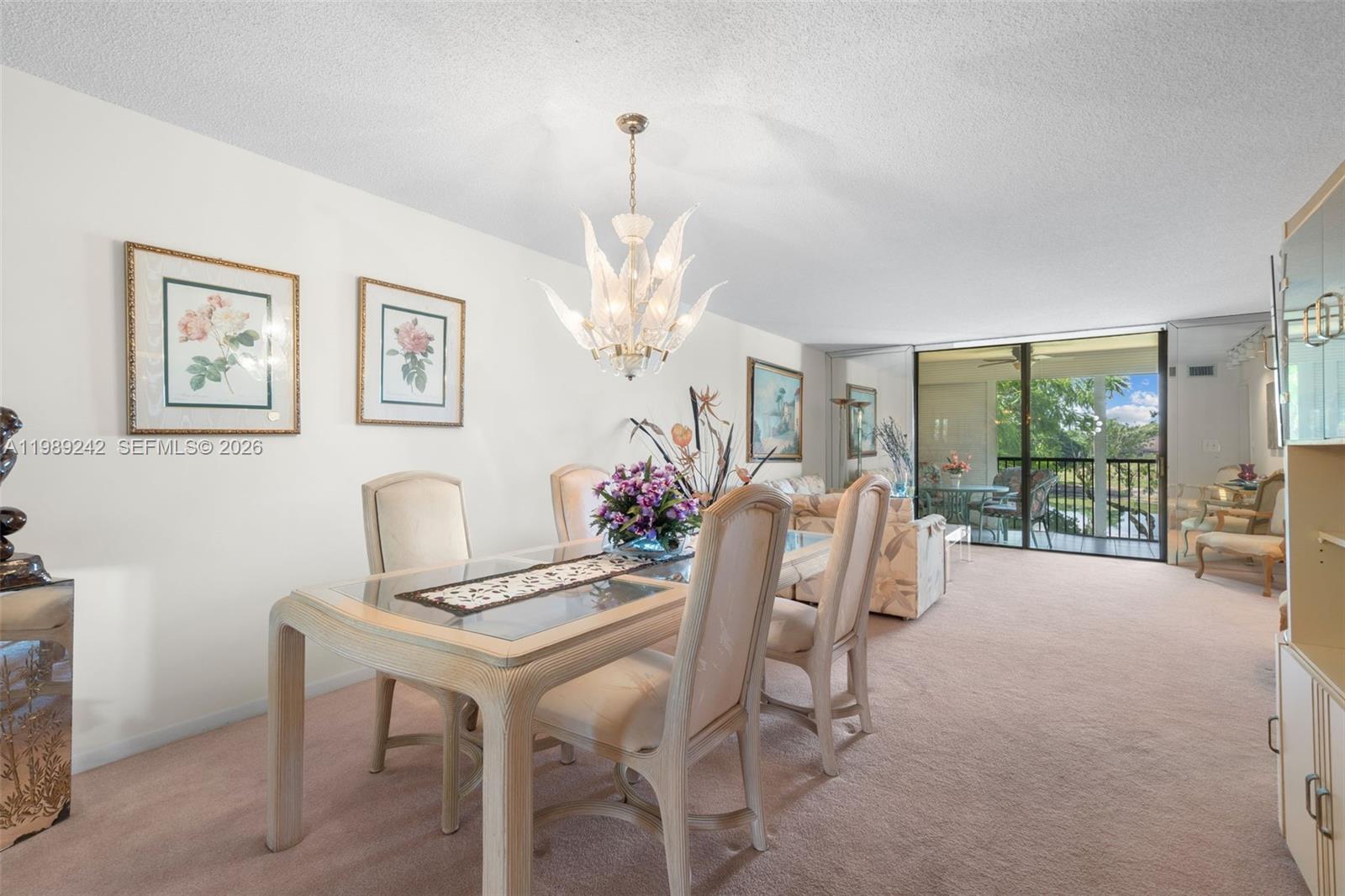 A dining area with a glass-topped table, chandelier, and carpeted floors leads to a balcony — 3485 Environ Blvd # C205, Lauderhill FL 33319, LAUDERHILL, FL
