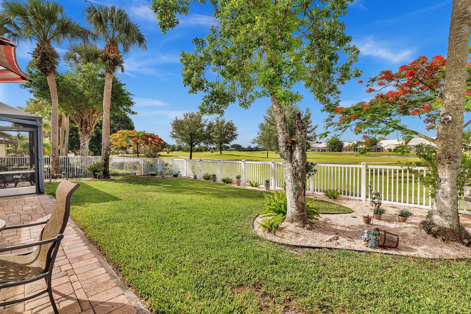 A backyard with a brick patio, green lawn, landscaping, and a white fence overlooking golf — 8099 Cassia Drive, Boynton Beach FL 33472, BOYNTON BEACH, FL